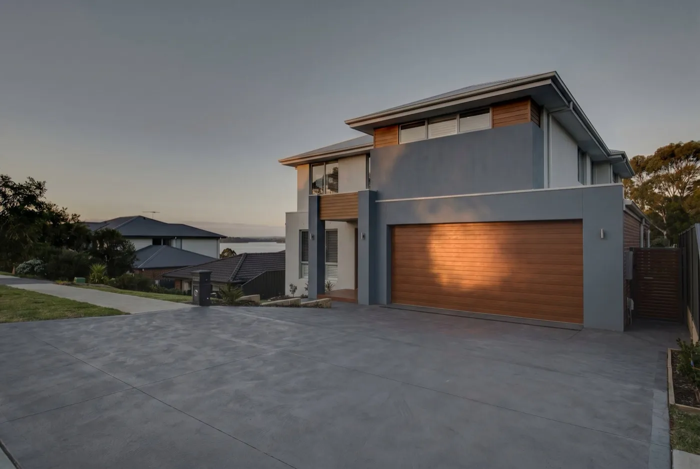 A steep driveway rising to a modern home with a wide sectional garage door, lake glimpse behind